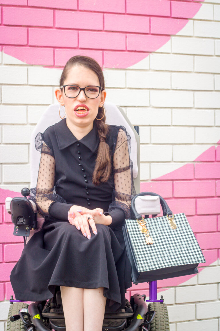 Amy is wearing a black dress and smiling to the camera. She is sitting in her wheelchair in front of a pink and white wall.