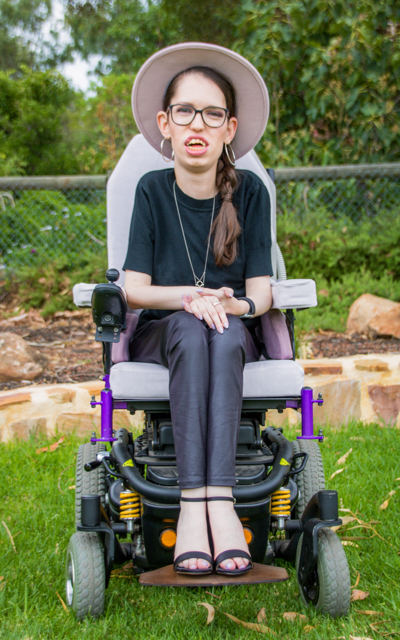 Amy is sitting in her wheelchair in a garden setting. She is wearing a purple felt hat, black knit top, leather pants and black high heels.